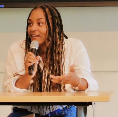 A woman sits at a table and speaks into a microphone.